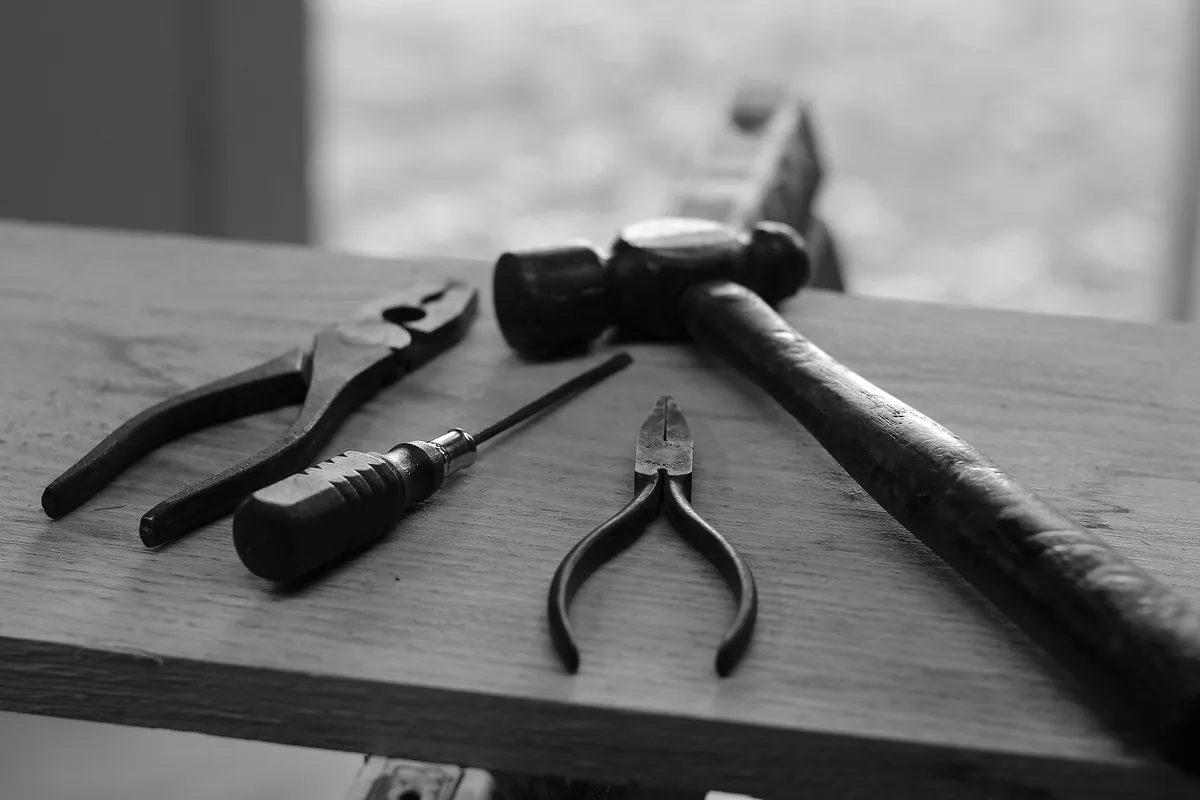 Illustrative black-and-white stock photo of pliers, a screwdriver and a hammer resting on a wooden workbench.
